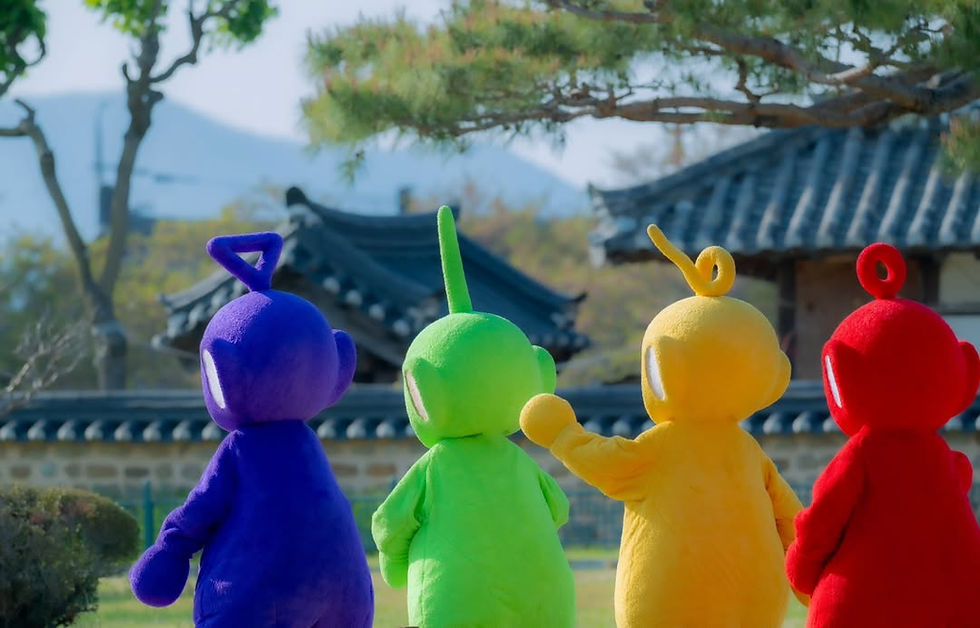 Four colorful characters, purple, green, yellow, and red, stand outside in front of a traditional building, with trees and mountains in the background.