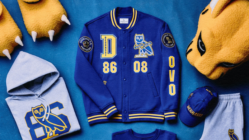 Flat lay of blue-and-gold Degrassi OVO apparel—jacket, hoodies, jerseys, pants, cap, tote, and plush paws.