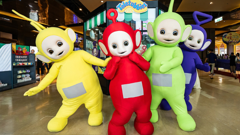 Four colorful Teletubbies mascots pose and smile in a mall beneath a Teletubbies sign, bright and playful.