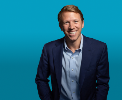 A professional headshot of a man in a blue shirt and dark blue suit jacket, photographed against a bright blue background.