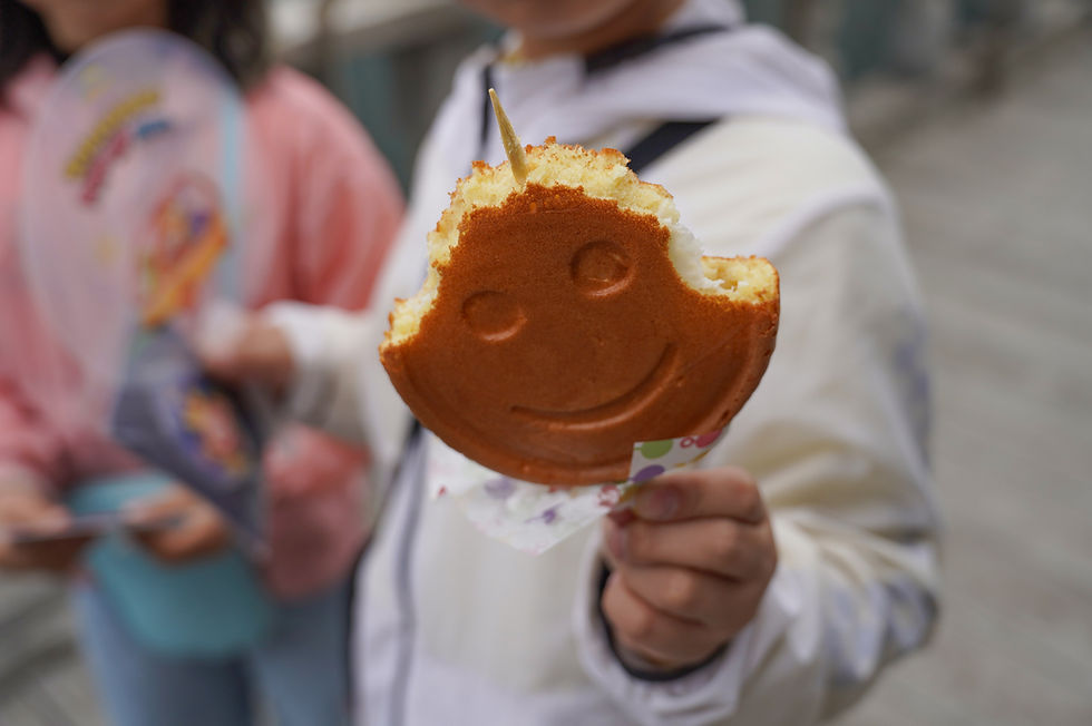 A person holds a "Tubby Toast" smiling pancake with a bite taken out. Another person holds a colorful fan in the background. The mood is playful.