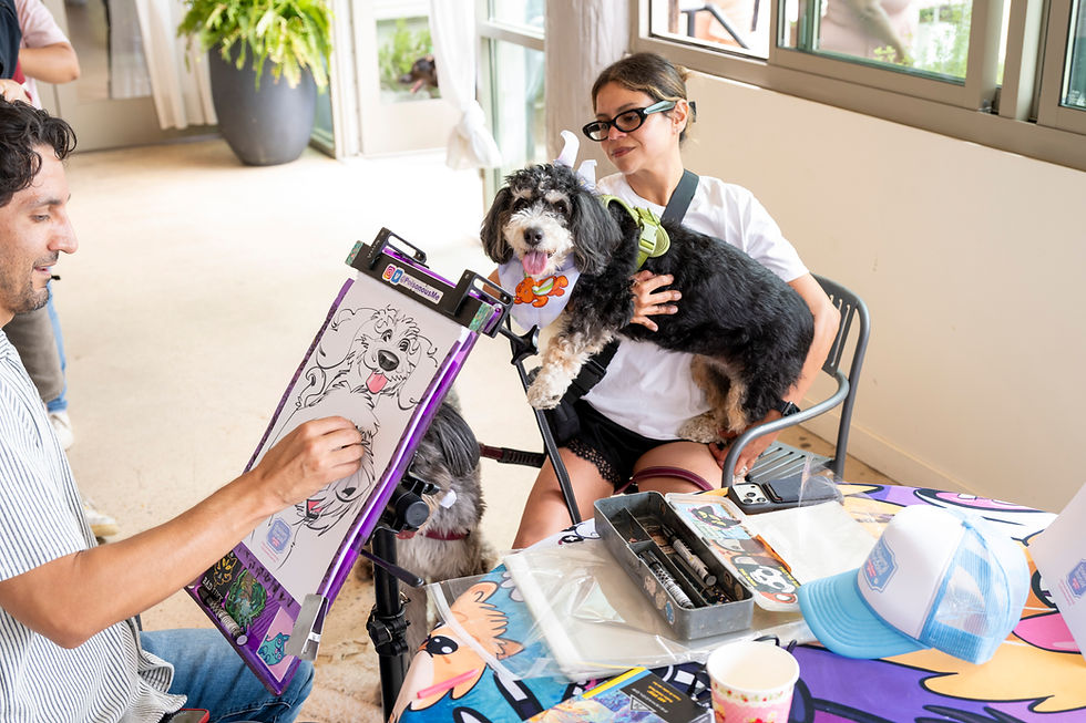 Woman holds a black dog while a man sketches its portrait indoors; a @poisonousMe sticker and art supplies sit on the table.