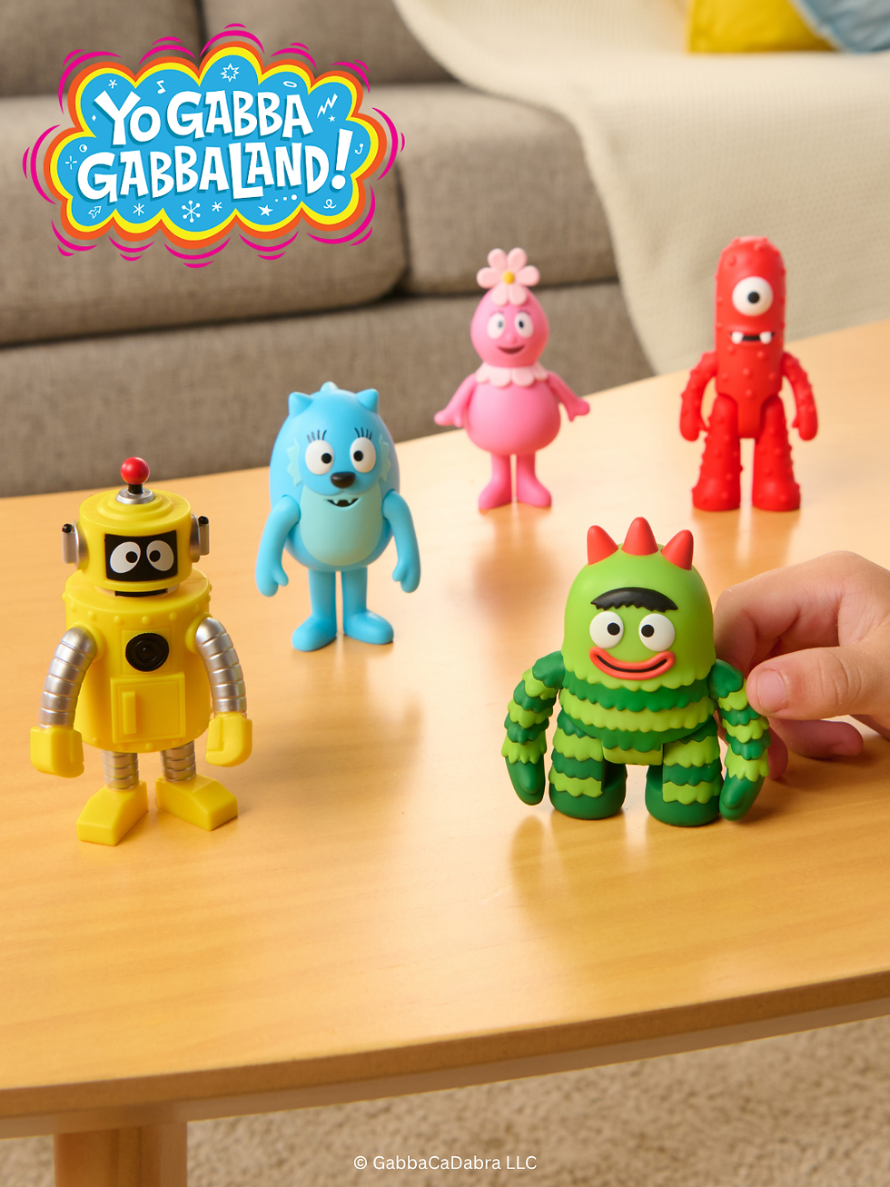 Colorful toy figures on a table. A hand holds a green figure. Yo Gabba Gabba! logo above.