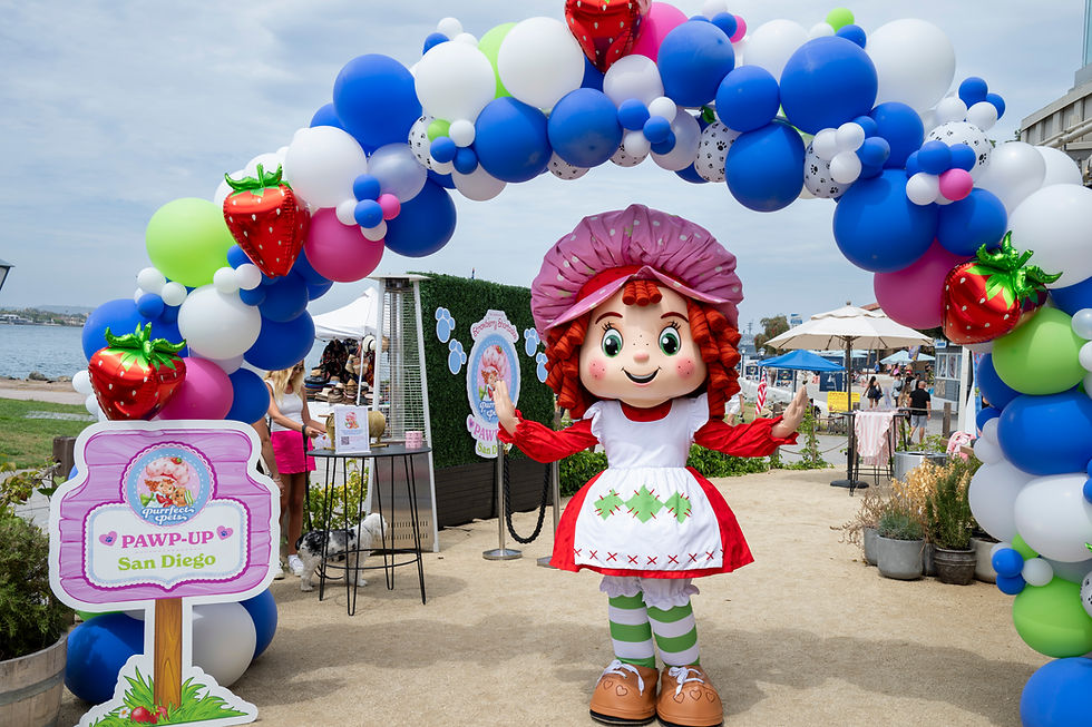 Strawberry Shortcake mascot under a blue-white balloon arch at a waterfront event; sign reads Pawp-Up San Diego.