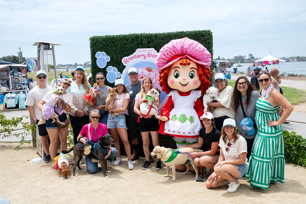 Group poses with dogs beside a Strawberry Shortcake mascot and sign at a sunny waterfront event.