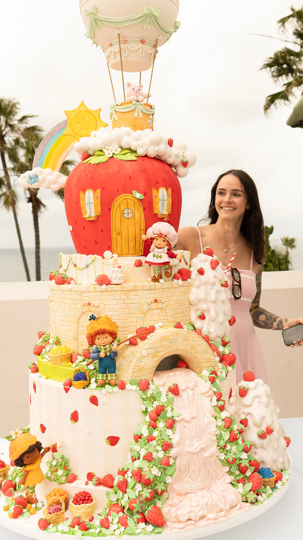 Cake Artist Ashley Holt smiles beside a giant Strawberry Shortcake fantasy cake with rainbow, dolls and hot-air balloon on a beach terrace