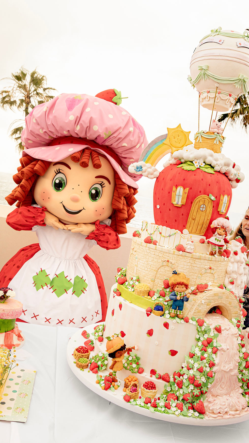 Smiling Strawberry Shortcake mascot beside a giant strawberry cake with tiny figures, rainbow, and hot-air balloon outdoors.