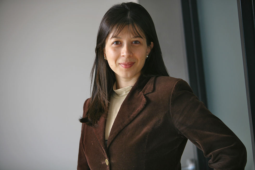 a woman in a beige shirt and brown jacket poses in front of a white wall