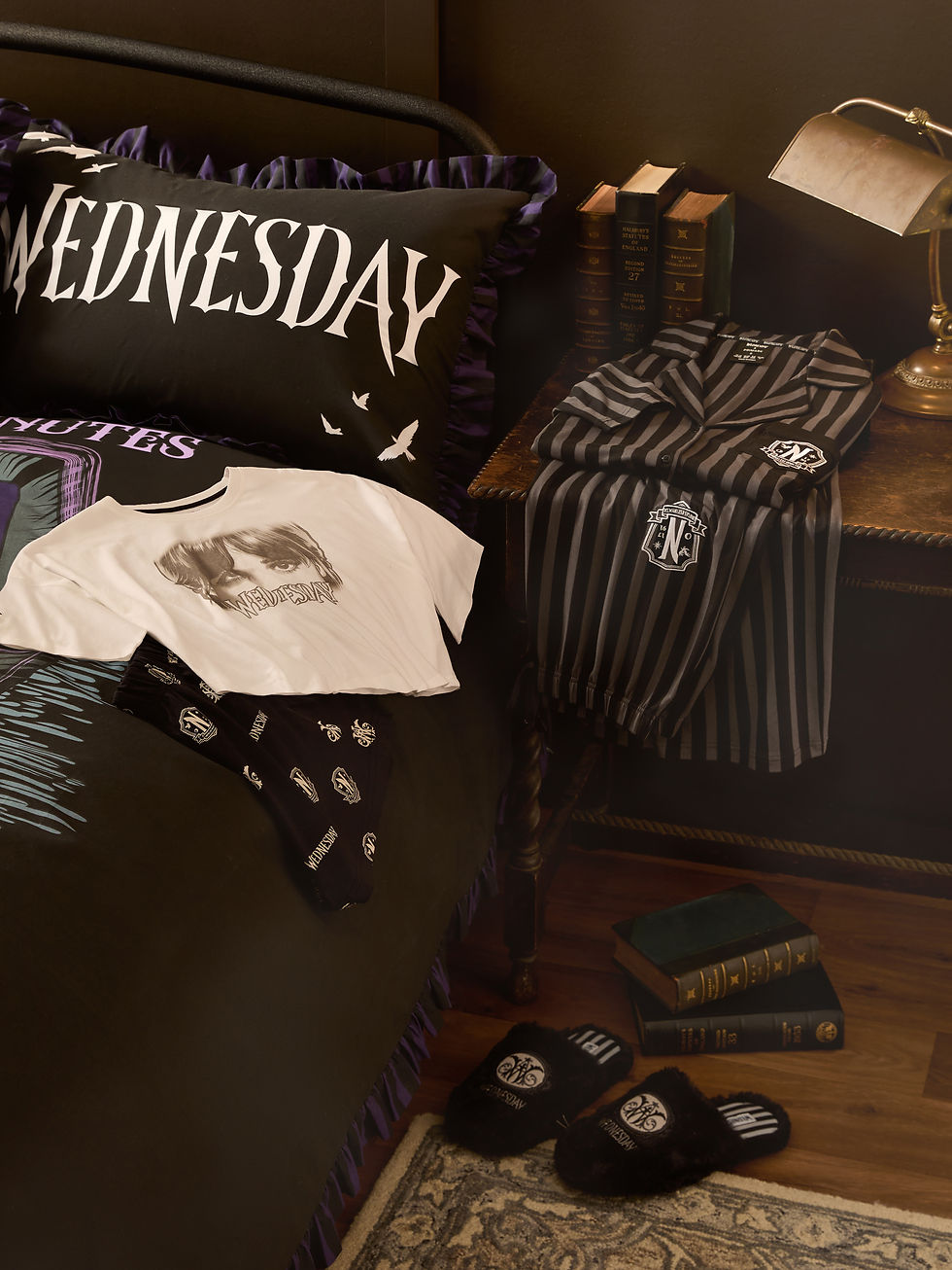 A collection of clothes from the Wednesday x Primark collection. A bed is covered in black Wednesday branded bedsheets. On top of the bed is a white Wednesday branded t-shirt and black Wednesday branded trousers. On top of the desk to the right is a set of striped wednesday branded pajamas . There are also Wednesday branded slippers on the floor.