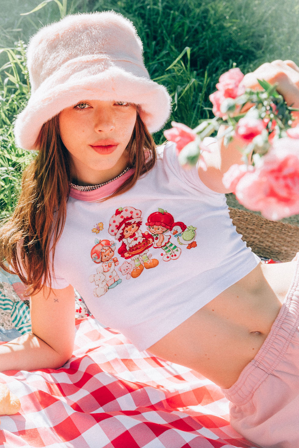 A teen lying on a red-checkered blanket, wearing a pink fuzzy hat and a white shirt with pink trim and strawberry-themed cartoon characters.