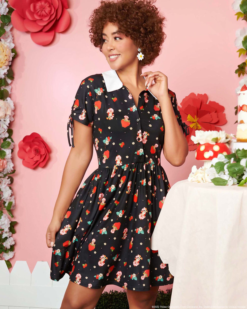 A woman with short curly brown hair wearing a vintage-style dress with a pattern of strawberries and a strawberry-themed character.
