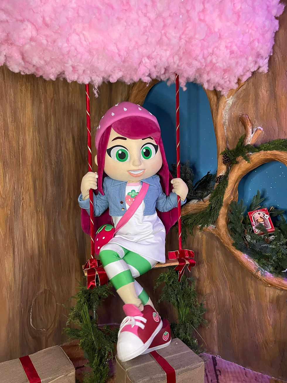 A strawberry-themed costumed character sits on a decorative swing in a holiday-themed wood cottage.