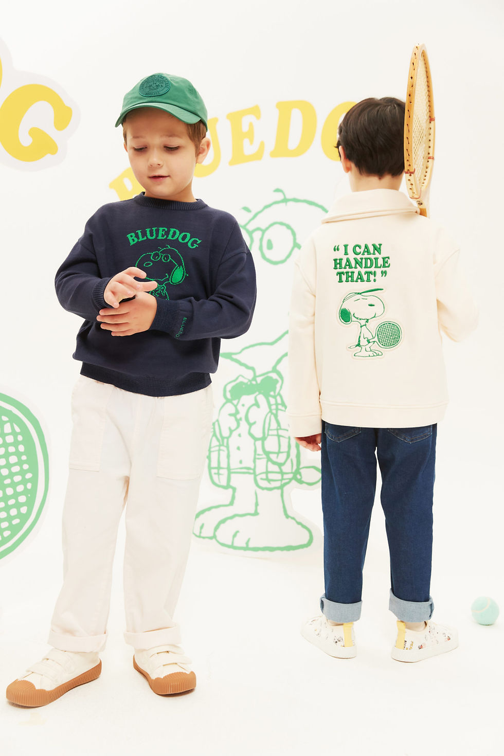 A boy faces forward, wearing a green hat and a dark pullover sweater that says "Bluedog" featuring a cartoon beagle. A boy faces backwards beside him, wearing a jacket that says "I can handle that" featuring the same cartoon beagle holding a racket.