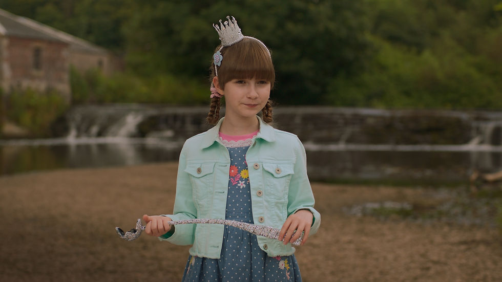 A young girl in a blue jean jacket and dress wears a small crown and holds a sparkly sceptre.