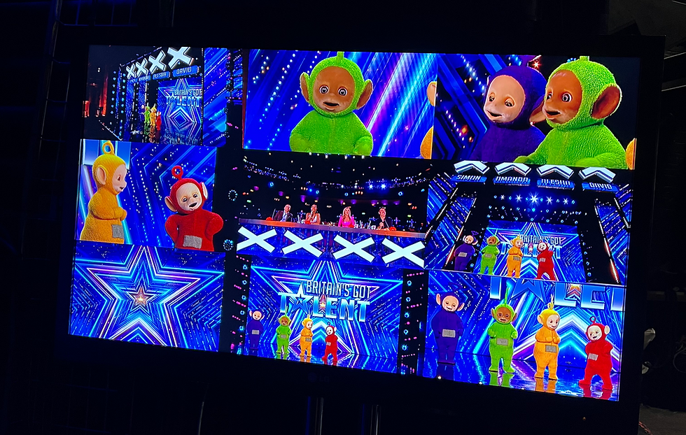 A backstage video screen displaying multiple camera angles of four alien costumed characters performing onstage in front of judges.