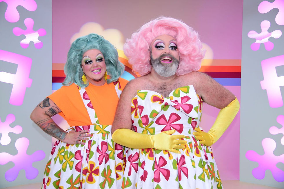 Two drag queens stand smiling in front of a pink background