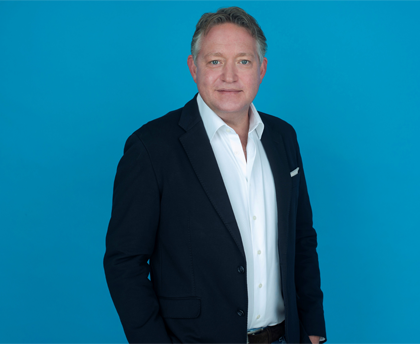 A headshot of a man in a suit standing and smiling in front of a blue background.