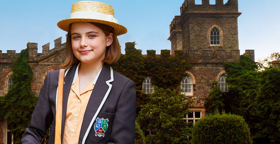 A fair-skinned girl, with short brown hair, standing in front of a castle. She is wearing a school uniform and a straw hat.