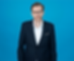 A headshot of a man in glasses and a suit, standing in front of a blue background.