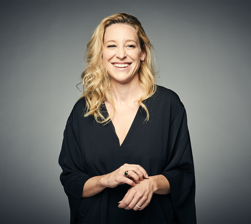 Headshot of a woman smiling with blonde hair, wearing a black blouse on a grey gradient background.