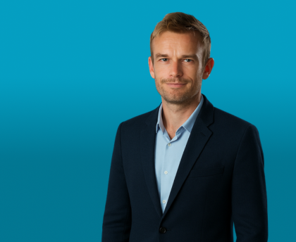 A professional headshot of a man wearing a dark blazer and light blue shirt, photographed against a bright blue background.