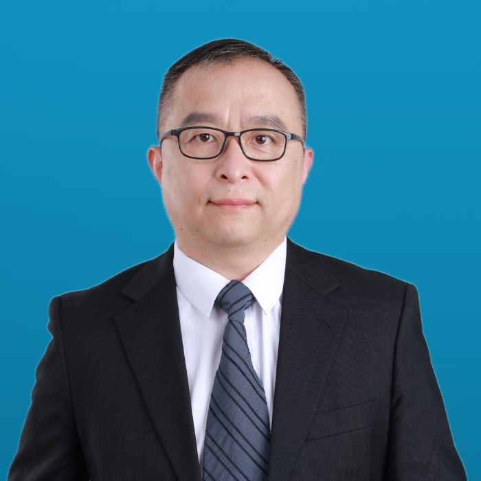 Headshot of a man with glasses in black blazer, white dress shirt and dark blue striped tie on a blue background. 