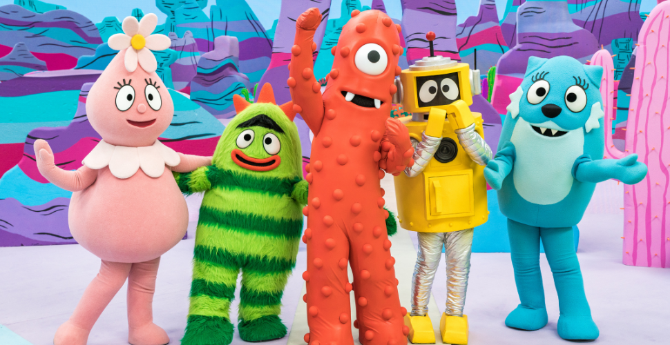 Five main Yo Gabba Gabba characters standing and posing with a woman dressed in purple overalls. They are in a colourful set mimicking a garden.