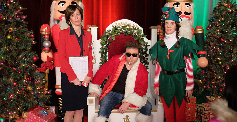 a man pretending to be Santa sitting next to a woman in red and an elf
